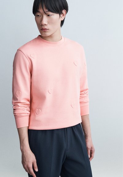 Jeune homme portant un sweat-shirt rose avec des cœurs en relief et un pantalon noir, regardant vers sa gauche sur un fond uni.