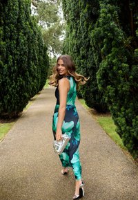 Eine Frau mit langen, welligen Haaren lächelt, während sie einen Gartenweg entlanggeht. Sie trägt ein Blumenkleid und hält eine silberne Clutch. Hohe grüne Hecken säumen den Gehweg.