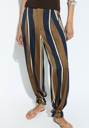 Femme portant un pantalon ample rayé marron, marine et crème avec des ourlets noués, debout pieds nus en tongs sur un fond uni.