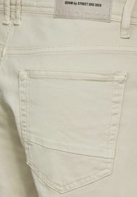 Bílé džínové kalhoty s zadní kapsou, která má vyšívané švy. Na štítku je napsáno "DENIM by STREET ONE MEN." Hladká textura.