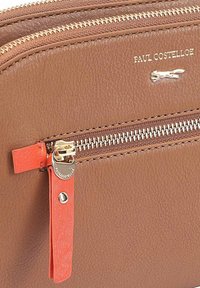 Bruine leren tas met textuur en gouden rits. Bevat een oranje accentlabel met een logo, aangevuld met een subtiele merknnaam die op het oppervlak is geperst.