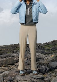 Ljusblå vattentät jacka över en grå mellanlager, i kombination med beige byxor och blågrå vandringsskor. Omgiven av stenig terräng.