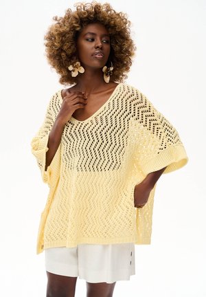 Kobieta z kręconymi włosami, nosząca duże kwiatowe kolczyki, luźny żółty sweter i białe szorty, pozująca z jedną ręką blisko szyi.