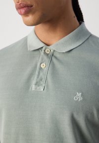 Gros plan d'une personne portant un polo vert clair à col avec deux boutons et de petites lettres blanches brodées M, O, P sur la poitrine.