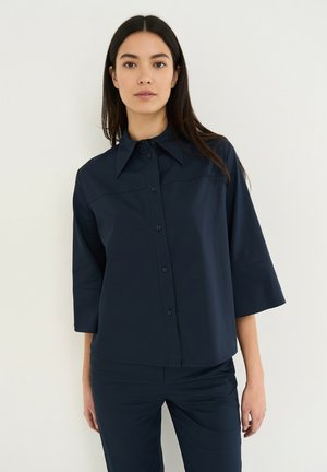 Femme aux longs cheveux foncés portant une chemise bleu marine foncé à boutons avec des manches trois-quarts et un pantalon assorti, debout devant un fond blanc.