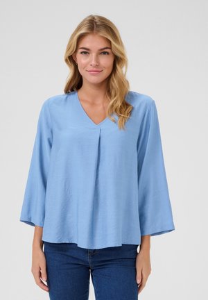 Blouse bleu clair, à col en V, avec des manches longues et une coupe ample. Fabriquée en tissu lisse avec une texture subtile, présentant un détail de pli central.