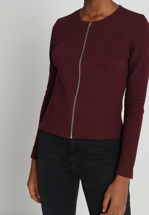 Femme portant un haut zippé texturé bordeaux à manches longues associé à un pantalon noir, torse et bras visibles sur un fond uni.