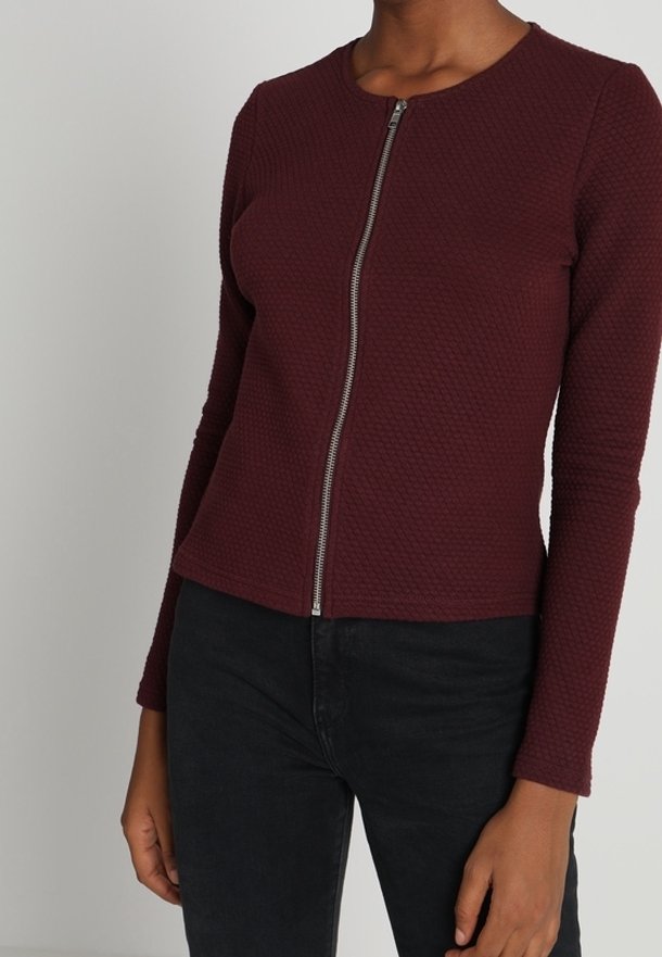 Femme portant un haut zippé texturé bordeaux à manches longues associé à un pantalon noir, torse et bras visibles sur un fond uni.