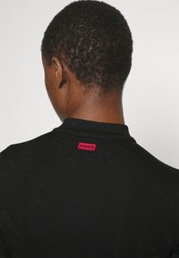 Chemise en maille noire présentant un motif texturé subtil, avec une étiquette logo rouge "HUGO" au niveau de l'encolure.