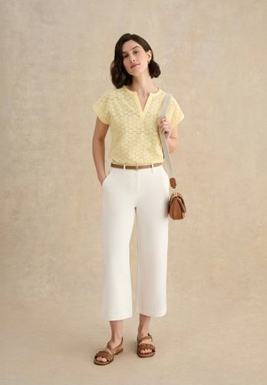 Vrouw met een geel kortemouwen top, witte cropped broek, bruine sandalen en een bruine crossbodytas, staand tegen een neutrale achtergrond.