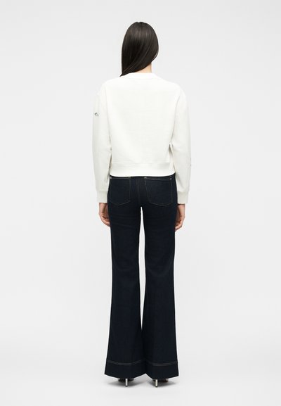 Γυναίκα που στέκεται με την πλάτη προς το μέρος μας, φοράει άσπρο μακρυμάνικο τοπ και σκούρα μπλε τζιν ψηλόμεσα καμπανέ με μαύρα τακούνια.