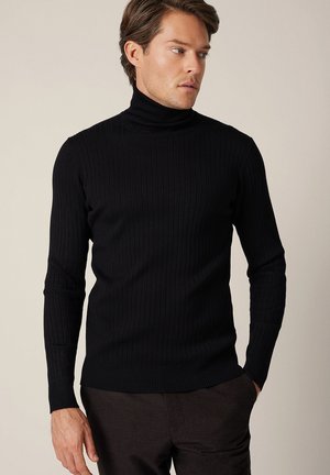 Mężczyzna w czarnym, prążkowanym golfie i ciemnych spodniach, patrzący na bok z jedną ręką w kieszeni na jednolitym tle.