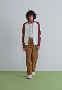 White zip-up jacket with maroon sleeves, paired with beige pants and white sneakers. Model stands on a green floor with a plain background.
