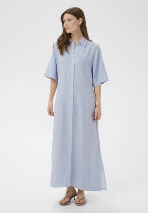 Femeie în picioare purtând o rochie maxi cu nasturi, cu dungi albastre și albe, mâneci scurte și sandale bej cu toc.