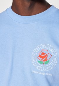 Světle modré bavlněné tričko s kruhovou grafikou červené růže, zeleným stonkem a textem "Visual Design Studio" níže v bílé barvě.