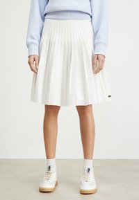 White pleated skirt with a high waist, made of lightweight fabric. Features a Lacoste logo accent and falls to mid-calf.