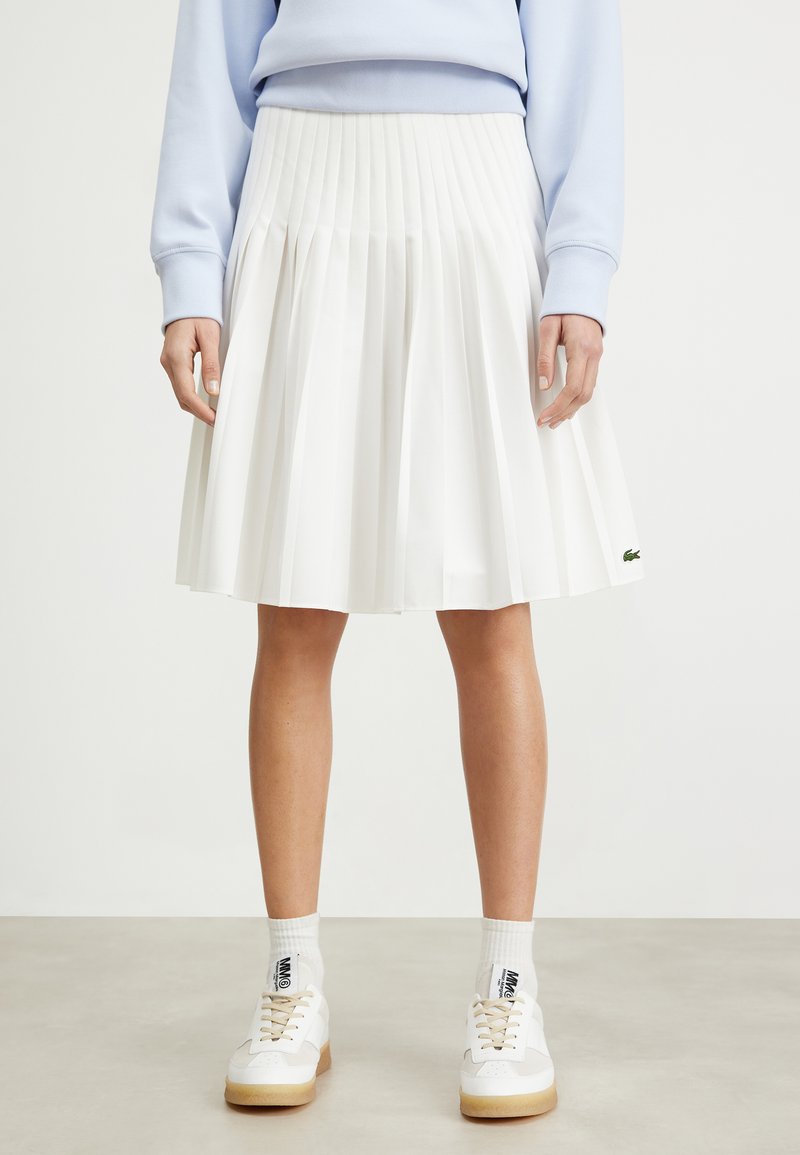 White pleated skirt with a high waist, made of lightweight fabric. Features a Lacoste logo accent and falls to mid-calf.