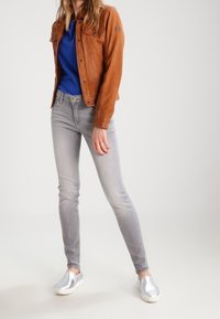 Brun läderjacka, blå topp, grå slim-fit jeans och silverfärgade slip-on skor. Funktioner inkluderar knappar, sömmar och släta texturer.