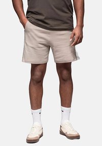 Shorts en coton beige, coupe droite, ceinture élastique, deux poches latérales. Porté avec un t-shirt noir, des chaussettes blanches et des baskets beiges.