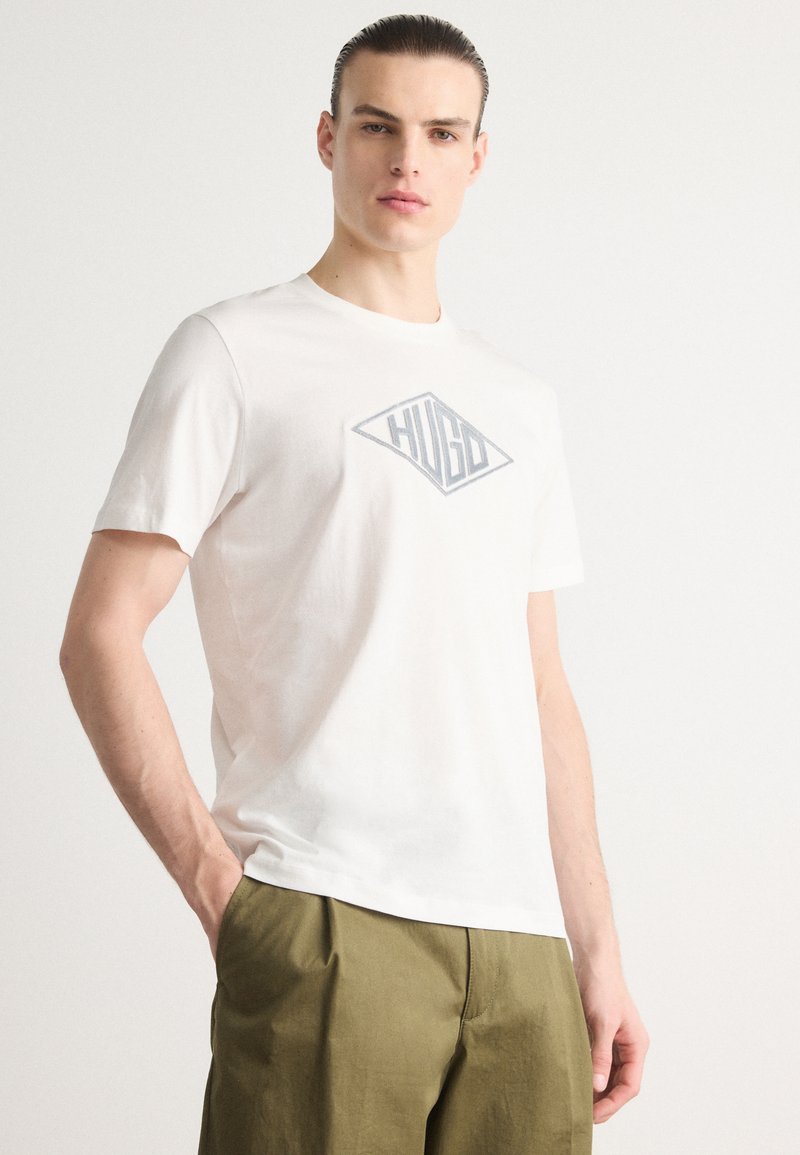 Hvit crew-neck t-skjorte laget av bomull, med et grått diamantlogo med "HUGO" i midten. Korte ermer og avslappet passform.