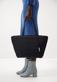 Czarna pleciona torba typu tote z dwoma uchwytami, o fakturowanej powierzchni i metce DKNY. Stylizowana z jasnoszarymi botkami za kostkę i niebieską sukienką.