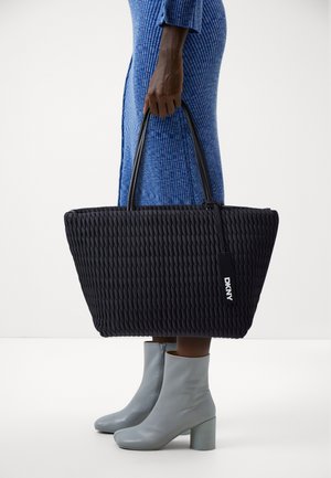 Borsa tote intrecciata nera con due manici, caratterizzata da una superficie testurizzata e un'etichetta DKNY. Abbinata a stivaletti grigio chiaro e un vestito blu.