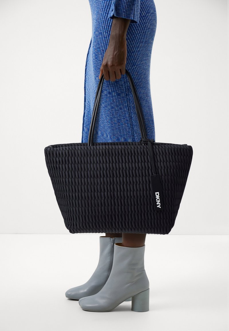 Czarna pleciona torba typu tote z dwoma uchwytami, o fakturowanej powierzchni i metce DKNY. Stylizowana z jasnoszarymi botkami za kostkę i niebieską sukienką.