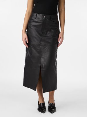 Mujer que lleva una falda midi de cuero negra con una abertura frontal, combinada con zapatos negros de punta afilada y una parte superior negra sin mangas.