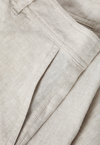 Beige linnen stof met een gestructureerd oppervlak, voorzien van gestikte naden en zakken, die een schone en natuurlijke esthetiek uitstraalt.