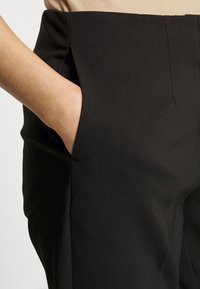 Svarta byxor med en slät textur och sidofickor, med skräddarsydda sömmar och en figurnära design, visas mot en neutral bakgrund.