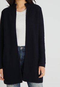 Námořnický pletený cardigan s vysokým límcem, dlouhými rukávy a otevřeným předním designem. Nositý přes lehký ribbovaný top a džíny.