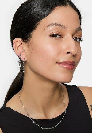 Boucles d'oreilles en argent en forme de créoles et un collier délicat en chaîne avec de petites accents, portés par une personne aux cheveux raides et foncés et portant un haut neutre.