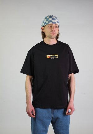 Junger Mann trägt ein schwarzes Grafik-T-Shirt mit Autoschriftzug, blaue Jeans und eine blau-weiß karierte Strickmütze, steht vor einem einfarbigen Hintergrund.