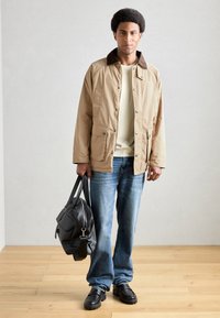 Beige jakke med brun krage, kremfarget genser under, blå jeans, svarte sko, holder en sort lærveske, står på tregulv.