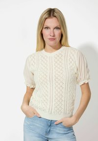 Dicker cremefarbener Pullover mit Rautenmuster, kurzen Puffärmeln und geripptem Saum. Getragen mit hellblauen Jeans. Weicher Stoff, leichtes Design.