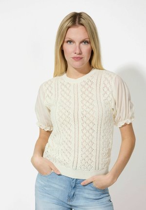 Pull en tricot crème avec un motif en losanges, manches courtes bouffantes et ourlet côtelé. Porté avec un jean bleu clair. Texture douce, design léger.