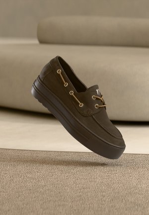 Mocassin décontracté marron foncé avec semelle épaisse et détails de lacets couleur fauve, flottant au-dessus d’un tapis beige texturé avec un canapé beige en arrière-plan.