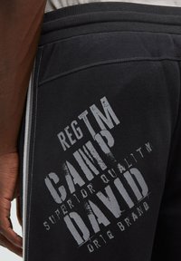 Schwarze Jogginghosen mit strukturiertem Finish, ausgestattet mit weißen Streifen und kräftig gedruckten Buchstaben in verschiedenen Größen im Bereich der Gesäßtasche.