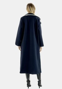 Langer marineblauer Mantel mit strukturiertem Design, ausgestattet mit weißem Kragen, weiten Ärmeln und glatter Textur. Kombiniert mit enganliegenden schwarzen Stiefeln.