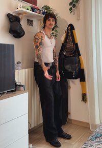 Sorte denimbukser med en avslappet passform, hvit tanktop og svarte sko. Har tatoveringer på armene og et sølvbelte. Veggdekorasjonen inkluderer planter.