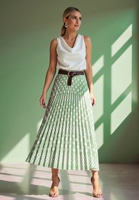 Maxi spódnica w jasnym zielonym kolorze w białe kropki, plisowany fason, zestawiona z białą bluzką na cowl neck i brązowym paskiem, na tle zielonego tła.