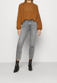 Pletený svetr v spáleně oranžové barvě s kabelovým vzorem, v kombinaci se šedými džíny s vysokým pasem. Modelka má na sobě černé kotníkové boty na podpatku.