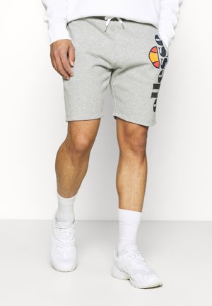 Mann trägt hellgraue Shorts mit großem schwarzem und orangefarbenem "ellesse"-Logo am linken Bein, weiße Socken und weiße Turnschuhe.