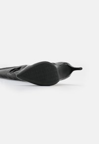 Zapato de tacón alto negro con punta afilada y textura de cuero suave; la suela presenta un patrón de marca en relieve; diseño elegante y minimalista.