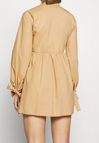 Abito corto di colore beige con vita arricciata, lunghe maniche a pallone con dettagli a lacci ai polsi e tessuto di cotone morbido. Vista posteriore mostrata.