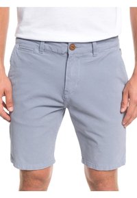 Muž v svetlo šedých neformálnych šortkách s predným gombíkom a zipsom, stojaci s uvoľnenými rukami pozdĺž tela na bielom pozadí.