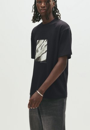 Black cotton t-shirt with a square graphic featuring abstract white and gray patterns, short sleeves, and a relaxed fit.