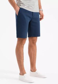 Marineblaues Shorts aus glattem Material, mit Fronttaschen und tailliertem Schnitt. Kombiniert mit weißen Slippers.