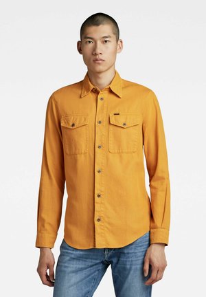 Camicia arancione a maniche lunghe con bottoni, due tasche sul petto, bottoni in metallo e orlo arrotondato, realizzata in tessuto liscio. Indossata con jeans blu.