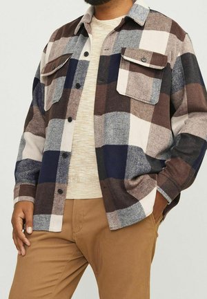 Mann trägt braunes, marineblaues, cremefarbenes und graues kariertes Hemd über einem beigen Pullover und hellbraune Hose, Hand in der Tasche.